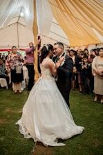 A couple is elegantly dressed and dancing closely in what appears to be a wedding reception. The woman is wearing a white wedding gown with intricate lace details, while the man is in a black suit with a boutonniere. They are surrounded by guests who are watching, taking photos, and smiling. The setting is a large tent with warm, golden drapery hanging from the ceiling, and the ground is covered with green grass.