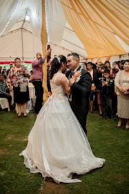 A couple is elegantly dressed and dancing closely in what appears to be a wedding reception. The woman is wearing a white wedding gown with intricate lace details, while the man is in a black suit with a boutonniere. They are surrounded by guests who are watching, taking photos, and smiling. The setting is a large tent with warm, golden drapery hanging from the ceiling, and the ground is covered with green grass.
