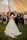 A couple is elegantly dressed and dancing closely in what appears to be a wedding reception. The woman is wearing a white wedding gown with intricate lace details, while the man is in a black suit with a boutonniere. They are surrounded by guests who are watching, taking photos, and smiling. The setting is a large tent with warm, golden drapery hanging from the ceiling, and the ground is covered with green grass.