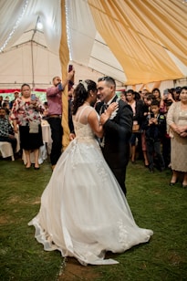 A couple is elegantly dressed and dancing closely in what appears to be a wedding reception. The woman is wearing a white wedding gown with intricate lace details, while the man is in a black suit with a boutonniere. They are surrounded by guests who are watching, taking photos, and smiling. The setting is a large tent with warm, golden drapery hanging from the ceiling, and the ground is covered with green grass.