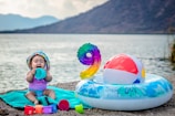 A baby wearing a colorful hat and swimsuit is sitting on a turquoise towel near a lake. The baby is surrounded by various toys, including colorful cups and a small beach ball. Nearby, there is an inflatable ring with flower patterns and a large multicolored number nine balloon. A serene lake and mountains are in the background.