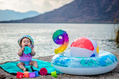 A baby wearing a colorful hat and swimsuit is sitting on a turquoise towel near a lake. The baby is surrounded by various toys, including colorful cups and a small beach ball. Nearby, there is an inflatable ring with flower patterns and a large multicolored number nine balloon. A serene lake and mountains are in the background.