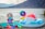 A baby wearing a colorful hat and swimsuit is sitting on a turquoise towel near a lake. The baby is surrounded by various toys, including colorful cups and a small beach ball. Nearby, there is an inflatable ring with flower patterns and a large multicolored number nine balloon. A serene lake and mountains are in the background.