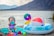A baby wearing a colorful hat and swimsuit is sitting on a turquoise towel near a lake. The baby is surrounded by various toys, including colorful cups and a small beach ball. Nearby, there is an inflatable ring with flower patterns and a large multicolored number nine balloon. A serene lake and mountains are in the background.