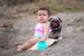 A small child in a pink and white swimsuit sits on sandy ground next to a blue container, with a pug dog wearing a black bandana sitting behind, licking its nose.