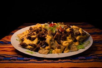 A plate filled with nachos topped with melted cheese, seasoned ground beef, lettuce, and diced tomatoes. The plate is placed on a colorful, striped tablecloth with a dark background.