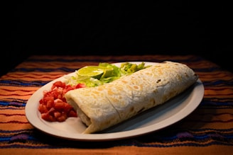 A delicious burrito is placed on a white plate, surrounded by a serving of chopped tomatoes, fresh shredded lettuce, slices of lime, and a small amount of guacamole. The plate rests on a vibrant, colorful tablecloth with orange, red, and blue stripes. The food presentation is appealing and appetizing.
