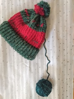 The image features a knitted beanie in red and green with a pom-pom on top, alongside a ball of yarn. The beanie appears to lie on a patterned surface, resembling birch bark with small dot details.