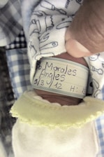 A close-up of a newborn baby's wrist with an identification bracelet. The bracelet displays handwritten details such as a name and date. The baby is wrapped in a soft, patterned fabric.