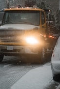 A large yellow tow truck is driving down a snowy road with its lights on. Snow covers the ground and the top of adjacent vehicles. The environment appears cold and overcast with snowflakes visible in the air.