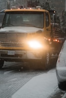 A large yellow tow truck is driving down a snowy road with its lights on. Snow covers the ground and the top of adjacent vehicles. The environment appears cold and overcast with snowflakes visible in the air.
