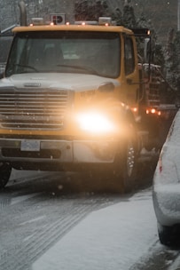 A large yellow tow truck is driving down a snowy road with its lights on. Snow covers the ground and the top of adjacent vehicles. The environment appears cold and overcast with snowflakes visible in the air.