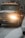 A large yellow tow truck is driving down a snowy road with its lights on. Snow covers the ground and the top of adjacent vehicles. The environment appears cold and overcast with snowflakes visible in the air.