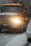 A large yellow tow truck is driving down a snowy road with its lights on. Snow covers the ground and the top of adjacent vehicles. The environment appears cold and overcast with snowflakes visible in the air.