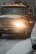 A large yellow tow truck is driving down a snowy road with its lights on. Snow covers the ground and the top of adjacent vehicles. The environment appears cold and overcast with snowflakes visible in the air.