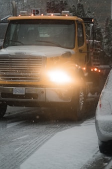 A large yellow tow truck is driving down a snowy road with its lights on. Snow covers the ground and the top of adjacent vehicles. The environment appears cold and overcast with snowflakes visible in the air.