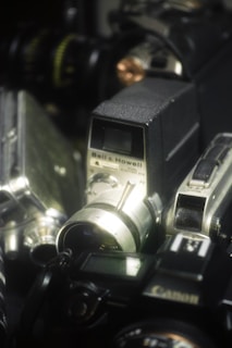 A collection of vintage cameras and equipment, including a Bell & Howell camera prominently displayed with a silver lens and black body. Other cameras are partially visible in the background, featuring various dials and metallic parts.