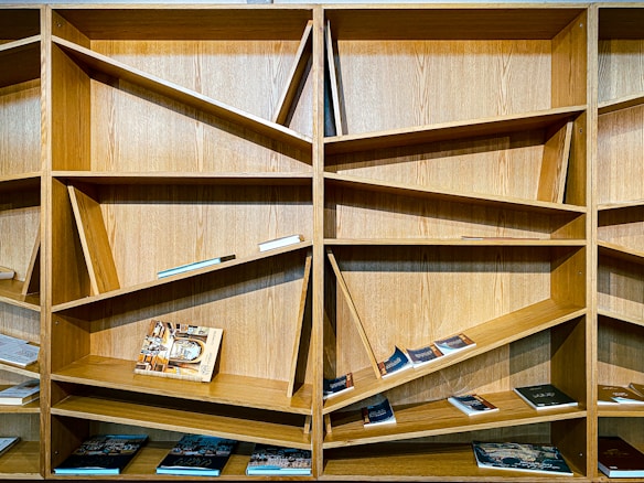 A wooden bookshelf with a unique and modern design features angular and slanted shelves. Several books are placed on the shelves, some laying flat while others are upright or slightly tilted.