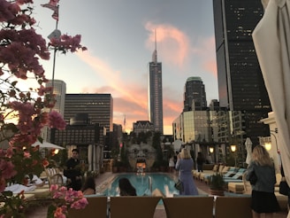 A rooftop pool area with modern buildings in the background, surrounded by lounge chairs and people relaxing. The sky is colorful with pink and orange hues from the sunset. Pink flowers are visible in the foreground, adding a touch of nature to the urban setting.