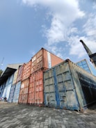 A stack of large, industrial shipping containers in various colors, including rusty orange, blue, and weathered steel, sits on a paved yard. The containers are placed next to a large industrial building with corrugated metal siding and an angled roof. The sky above is partly cloudy, offering a glimpse of blue amid the white clouds.