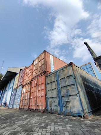 A stack of large, industrial shipping containers in various colors, including rusty orange, blue, and weathered steel, sits on a paved yard. The containers are placed next to a large industrial building with corrugated metal siding and an angled roof. The sky above is partly cloudy, offering a glimpse of blue amid the white clouds.