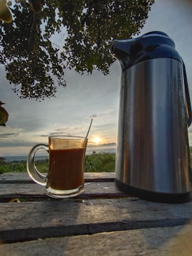 A glass mug filled with a hot beverage is resting on a wooden table outdoors. Next to it stands a large metallic thermos. The background features a scenic view of the sunset with a sky filled with clouds, and lush greenery is visible on the horizon. Overhead, tree branches with leaves partially frame the view.