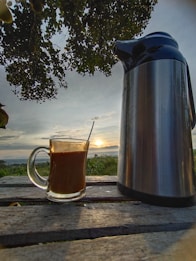 A glass mug filled with a hot beverage is resting on a wooden table outdoors. Next to it stands a large metallic thermos. The background features a scenic view of the sunset with a sky filled with clouds, and lush greenery is visible on the horizon. Overhead, tree branches with leaves partially frame the view.