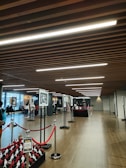 A modern exhibition hall with wooden flooring and ceiling elements, featuring rows of display panels with artwork. Red velvet ropes form barriers around a pedestal with small red and white sculptures. People are seen casually walking and observing the displays.