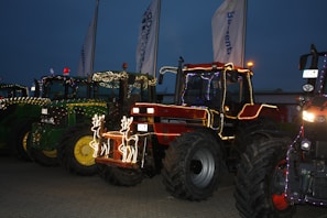 Several tractors are decorated with colorful string lights and festive adornments, placed in a row on a paved surface during dusk. Two illuminated reindeer figures are attached to the front of a red tractor.