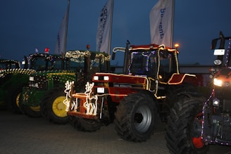 Several tractors are decorated with colorful string lights and festive adornments, placed in a row on a paved surface during dusk. Two illuminated reindeer figures are attached to the front of a red tractor.