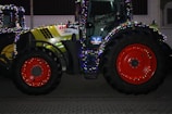A large agricultural tractor decorated with colorful string lights, featuring bright red wheels and a green and black body. It is parked on a paved surface, with another lit tractor partially visible in the background.