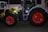 A large agricultural tractor decorated with colorful string lights, featuring bright red wheels and a green and black body. It is parked on a paved surface, with another lit tractor partially visible in the background.