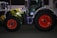 A large agricultural tractor decorated with colorful string lights, featuring bright red wheels and a green and black body. It is parked on a paved surface, with another lit tractor partially visible in the background.