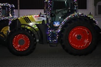 A large agricultural tractor decorated with colorful string lights, featuring bright red wheels and a green and black body. It is parked on a paved surface, with another lit tractor partially visible in the background.