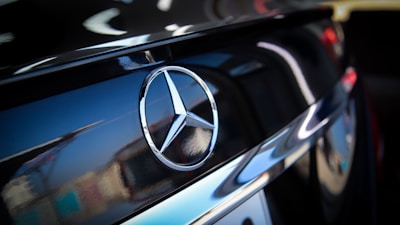 A close-up of a car displaying the Mercedes-Benz logo on a shiny black surface. The emblem, a three-pointed star, is prominently featured against a reflective background that captures some light and shadow effects.