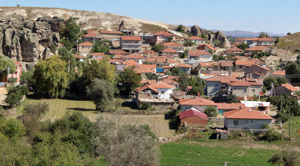 The image depicts a picturesque village nestled on a hillside, surrounded by lush greenery and trees. The houses in the village are painted in warm hues of red and orange, contrasting with the cool tones of the surrounding landscape. The perspective of the photo is from above, providing a bird's eye view of the village and its natural surroundings.