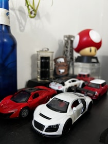 A collection of toy cars is displayed on a dark surface, including a white sports car in the foreground and a red sports car beside it. Other smaller toy vehicles are visible in the background. A blue bottle and a large red mushroom with white spots adorn the setup, along with a tower-like structure and other decorative elements.