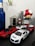 A collection of toy cars is displayed on a dark surface, including a white sports car in the foreground and a red sports car beside it. Other smaller toy vehicles are visible in the background. A blue bottle and a large red mushroom with white spots adorn the setup, along with a tower-like structure and other decorative elements.