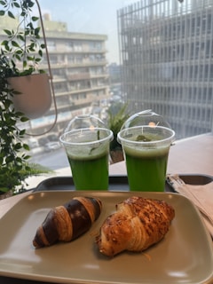 Two green beverages in plastic cups with dome lids are placed on a table with a view of urban buildings through a window. In front, there is a plate with two croissants, one of which appears to be chocolate-flavored. Several potted plants are visible, adding a touch of greenery.