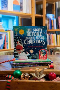 A festive children's book titled 'The Night Before The Night Before Christmas' by Kes Gray and Claire Powell is prominently displayed on a wooden table. The cover features a whimsical illustration of Santa Claus in the snow with vibrant colors, surrounded by Christmas-themed objects such as a shiny silver star, a red and green striped ornament, a spool of red thread, and other festive decorations. In the background, there is a well-stocked bookshelf filled with various books.