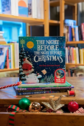 A festive children's book titled 'The Night Before The Night Before Christmas' by Kes Gray and Claire Powell is prominently displayed on a wooden table. The cover features a whimsical illustration of Santa Claus in the snow with vibrant colors, surrounded by Christmas-themed objects such as a shiny silver star, a red and green striped ornament, a spool of red thread, and other festive decorations. In the background, there is a well-stocked bookshelf filled with various books.
