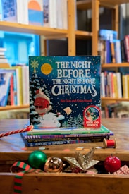 A festive children's book titled 'The Night Before The Night Before Christmas' by Kes Gray and Claire Powell is prominently displayed on a wooden table. The cover features a whimsical illustration of Santa Claus in the snow with vibrant colors, surrounded by Christmas-themed objects such as a shiny silver star, a red and green striped ornament, a spool of red thread, and other festive decorations. In the background, there is a well-stocked bookshelf filled with various books.