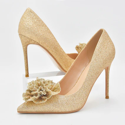 A close-up of a pair of blush pink Veloura heels resting elegantly on a soft ivory fabric background with subtle gold accents.