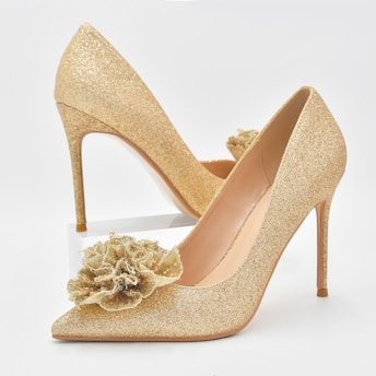 A pair of elegant high-heeled shoes with a shimmering gold finish and a decorative floral embellishment on the toe. The background is a plain white, emphasizing the luxurious texture and design of the shoes.