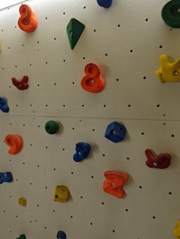 A climbing wall with various colorful holds made of plastic. The holds are attached to a beige wall with a grid of holes, allowing for adjustable positioning. The holds are in different shapes and colors, including blue, red, green, yellow, and orange.