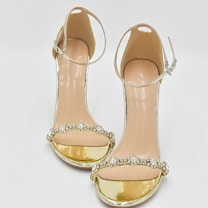 A pair of elegant high-heeled sandals designed with golden metallic bands and adorned with sparkling gemstone embellishments across the toes. The ankle strap is thin and has a buckle, providing additional support and style.