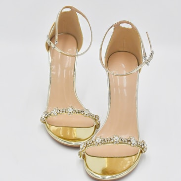 A pair of elegant high-heeled sandals designed with golden metallic bands and adorned with sparkling gemstone embellishments across the toes. The ankle strap is thin and has a buckle, providing additional support and style.
