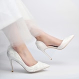 A pair of elegant white high-heeled shoes with floral embroidery are worn by a person. The shoes have a pointed toe design and the person's legs are slightly bent, wearing a sheer, delicate white fabric, possibly part of a dress.