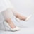 A pair of elegant white high-heeled shoes with floral embroidery are worn by a person. The shoes have a pointed toe design and the person's legs are slightly bent, wearing a sheer, delicate white fabric, possibly part of a dress.