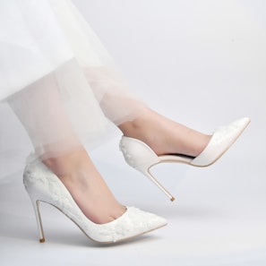 A pair of elegant white high-heeled shoes with floral embroidery are worn by a person. The shoes have a pointed toe design and the person's legs are slightly bent, wearing a sheer, delicate white fabric, possibly part of a dress.